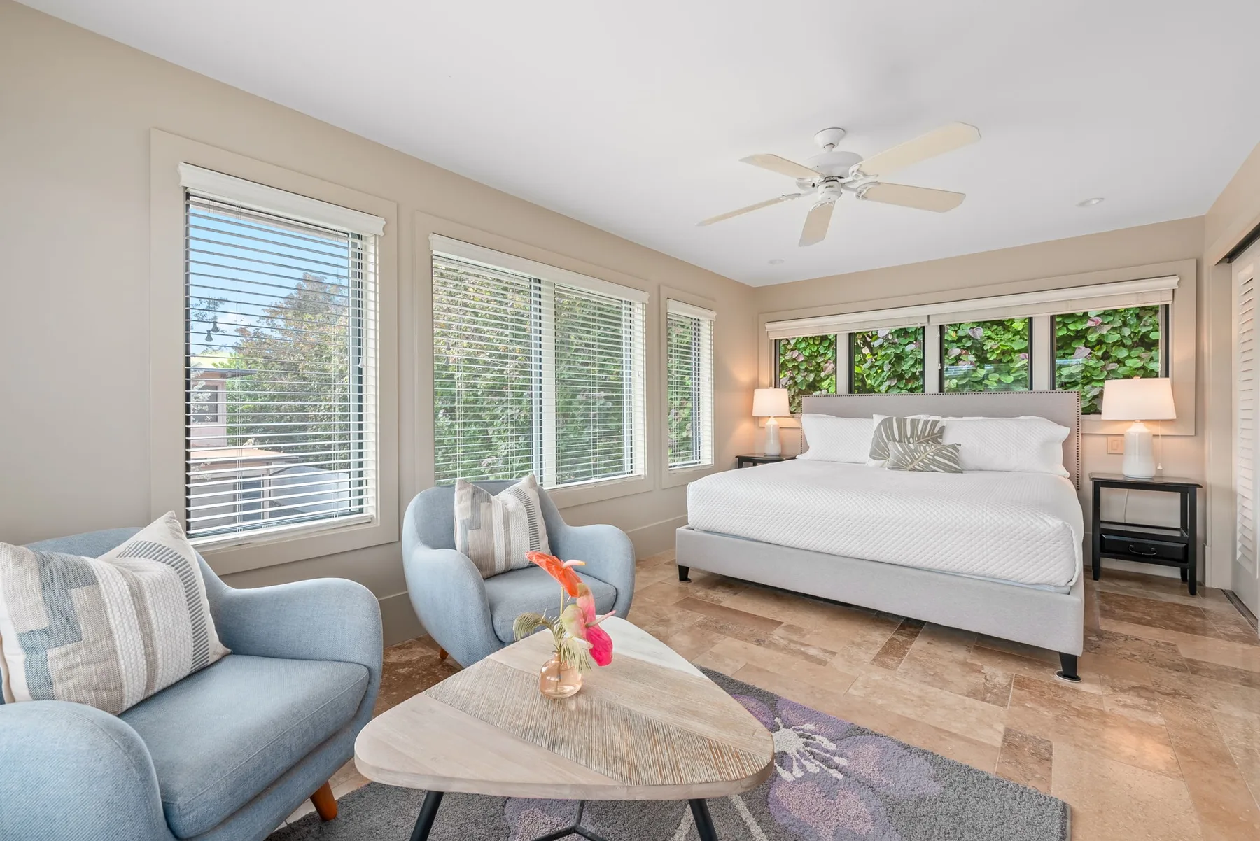 A bright bedroom has a large bed, blue seating, and windows overlooking greenery.