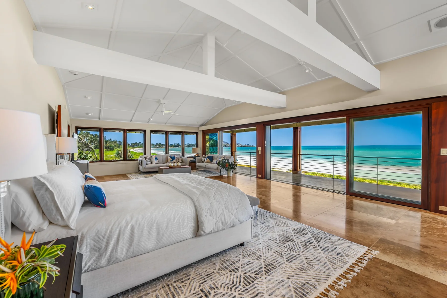A bright bedroom with a bed beside broad windows overlooking the ocean.