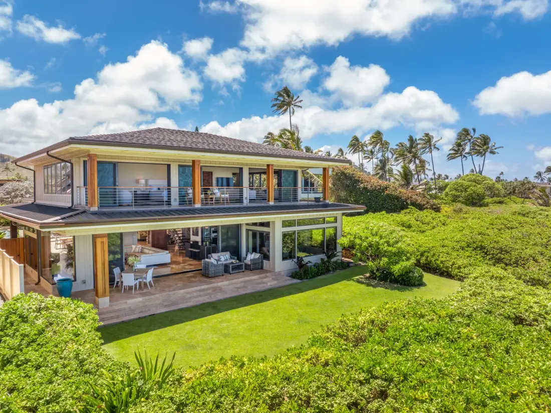 A two-story house with broad balconies stands beside a green lawn and tropical vegetation.