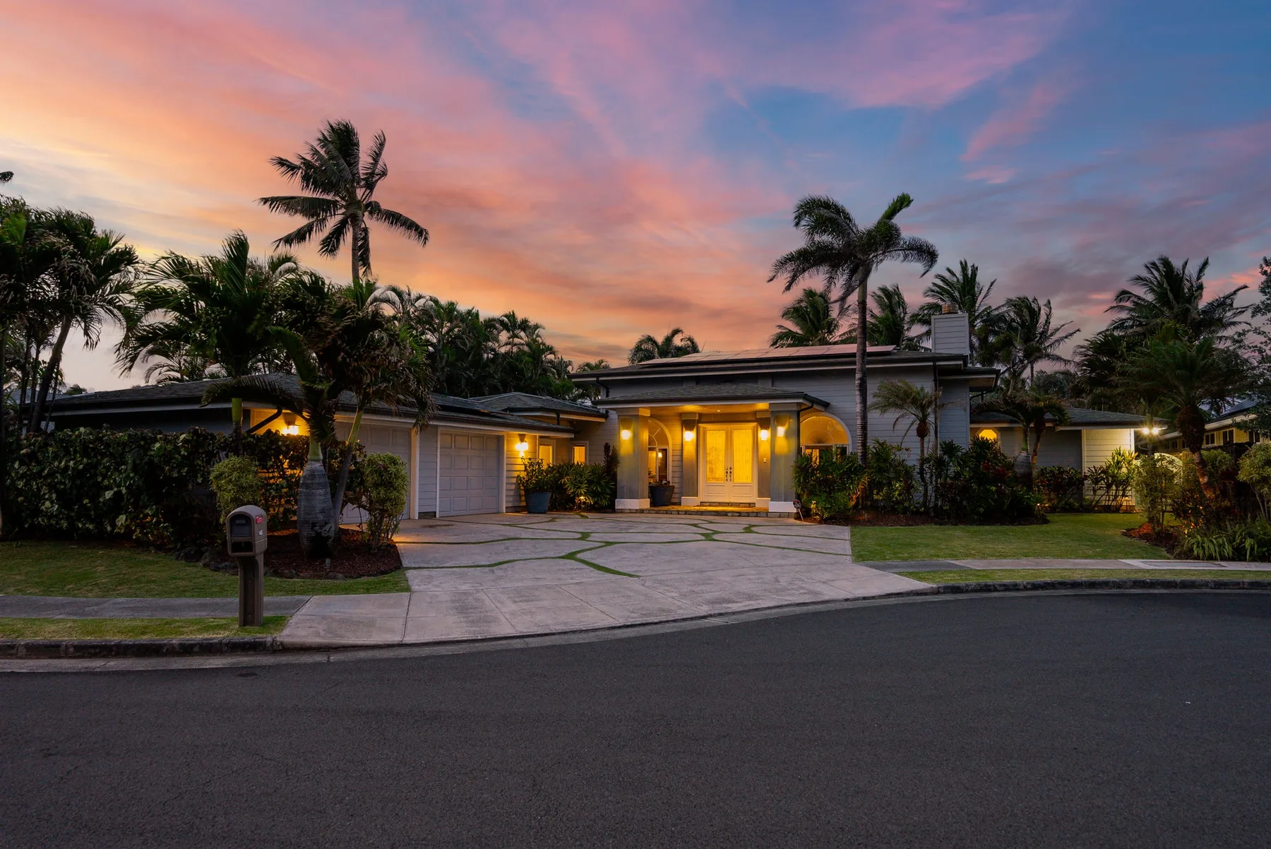 Una casa con palmeras y una entrada pavimentada bajo un cielo colorido al atardecer.