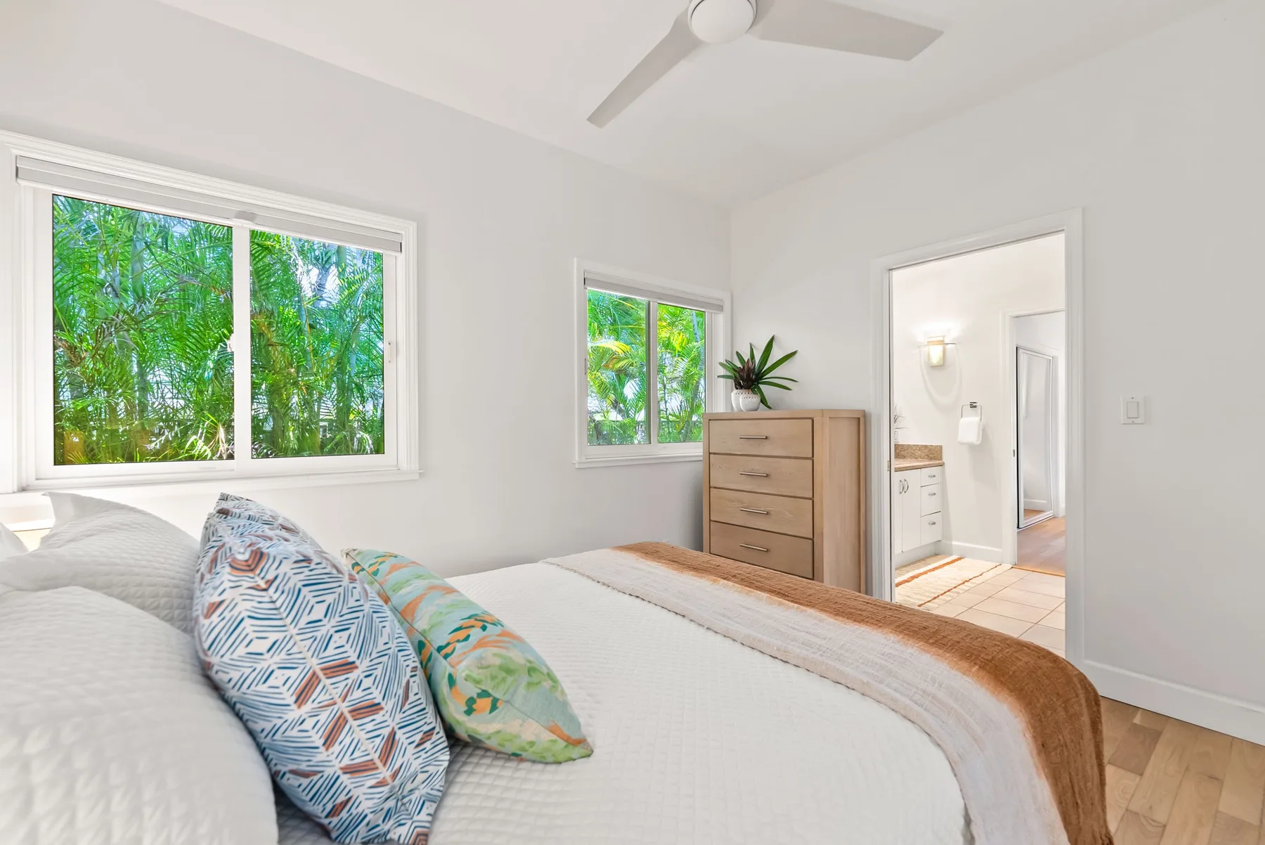 Un dormitorio luminoso con una cama, cojines estampados y ventanas con vistas a la vegetación.