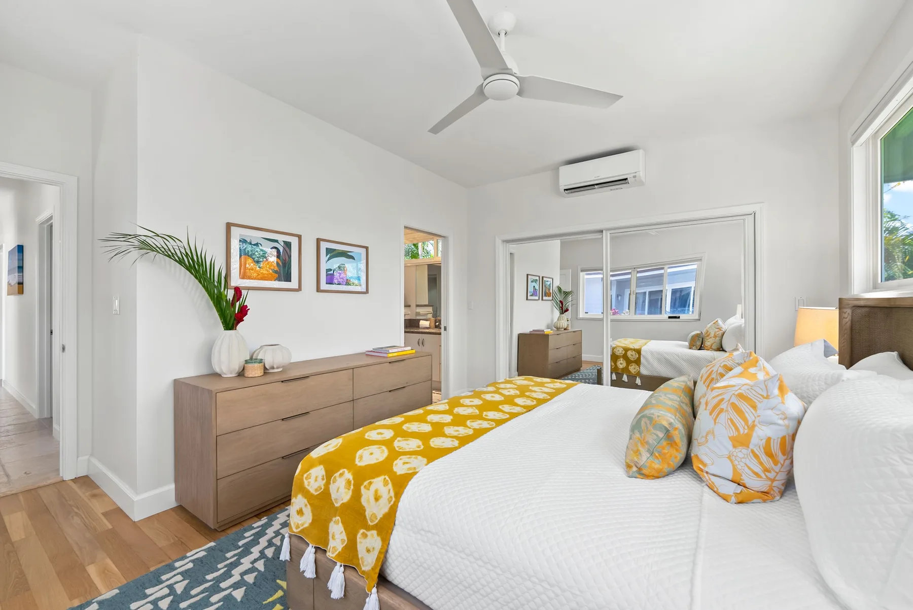 Un dormitorio luminoso con ropa de cama blanca, una manta amarilla estampada y un ventilador de techo.