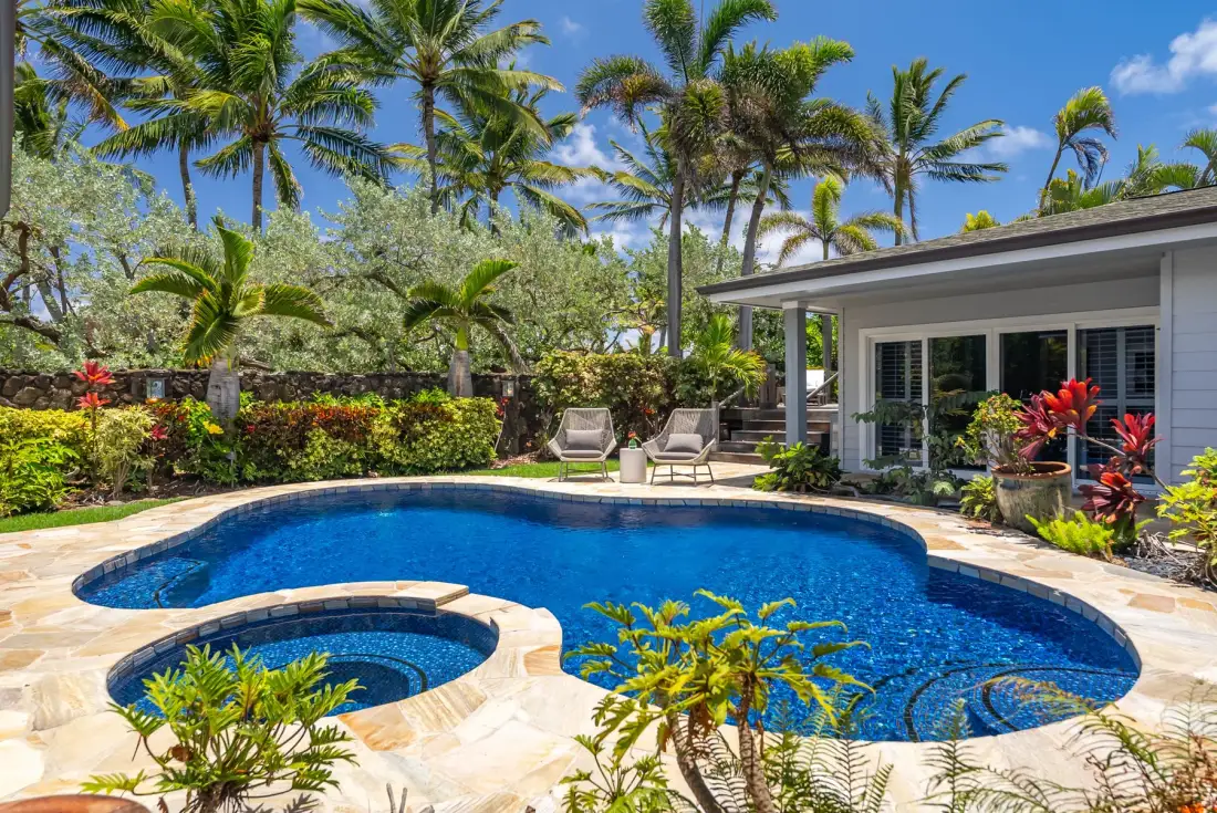 A curved swimming pool sits beside a patio with lounge seating and tropical plants.