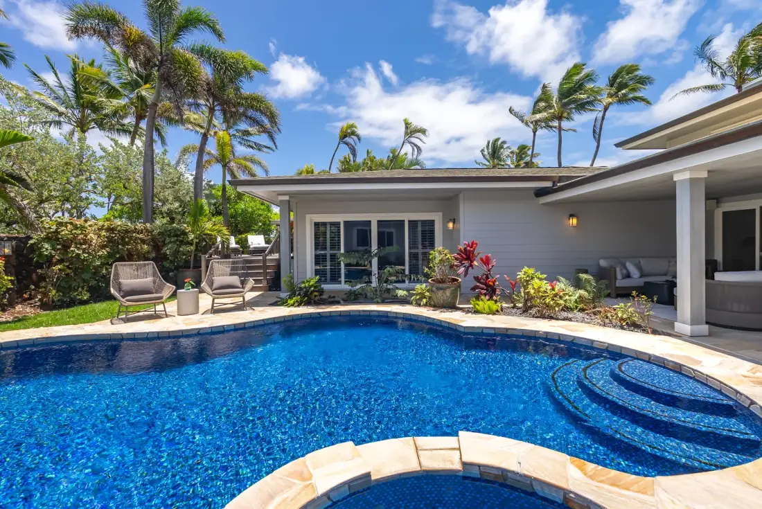 Piscina exterior junto a una casa moderna, con asientos en el patio y plantas tropicales.