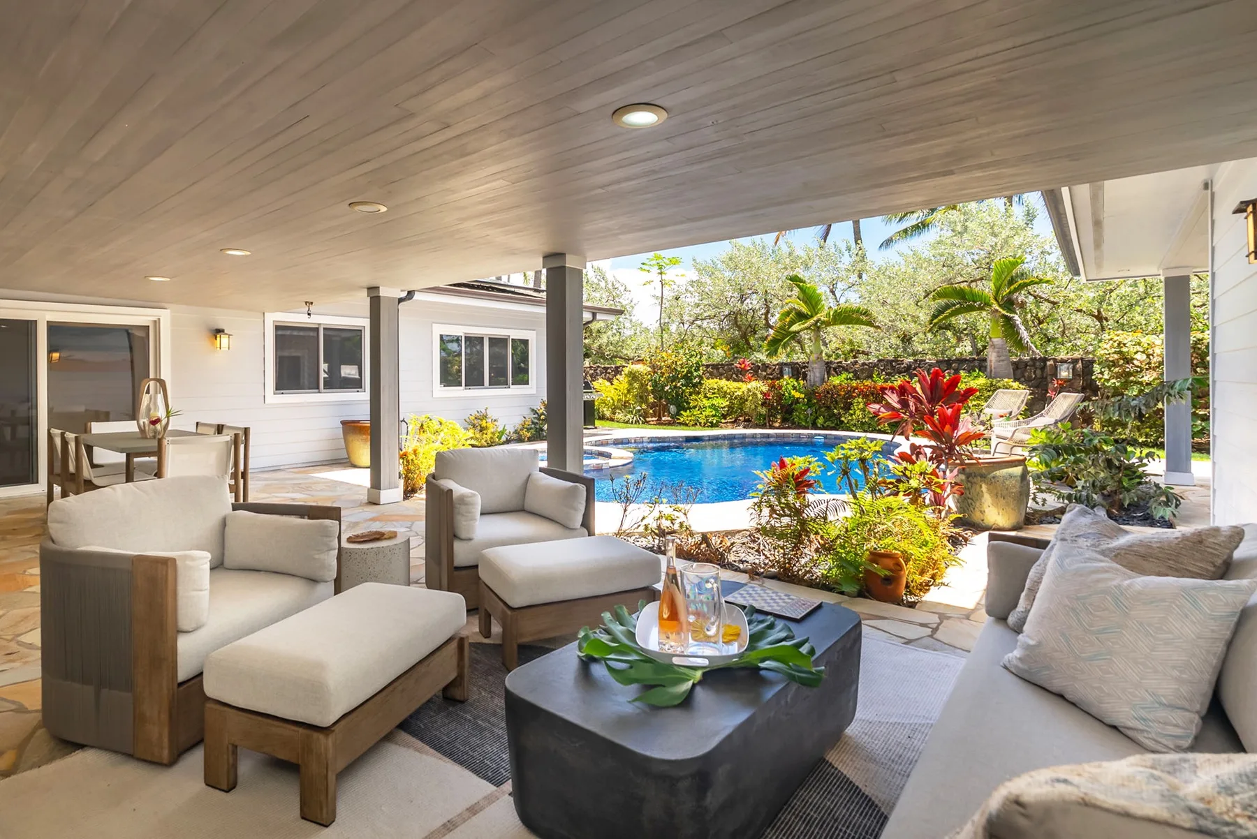 Una sala exterior cubierta con asientos acolchados da a una piscina y un jardín.