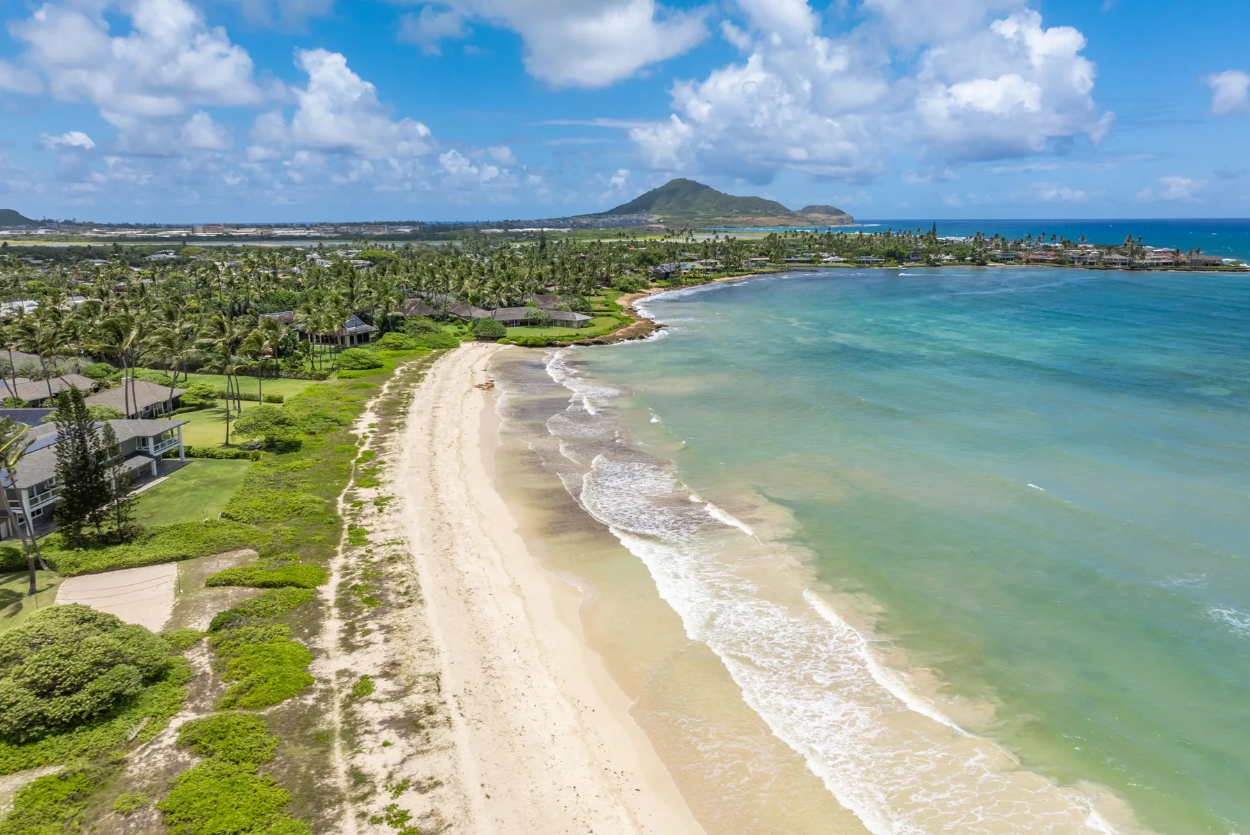 Vista aérea de una playa de arena curva junto a aguas turquesas y zonas verdes costeras.