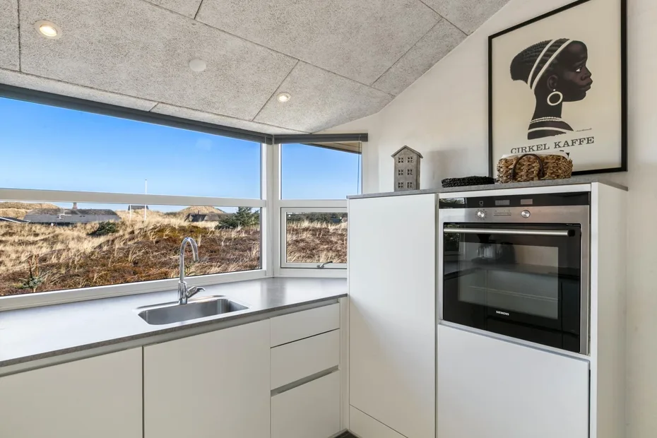 A modern kitchen with white cabinets, an oven, and wide windows overlooking the landscape.