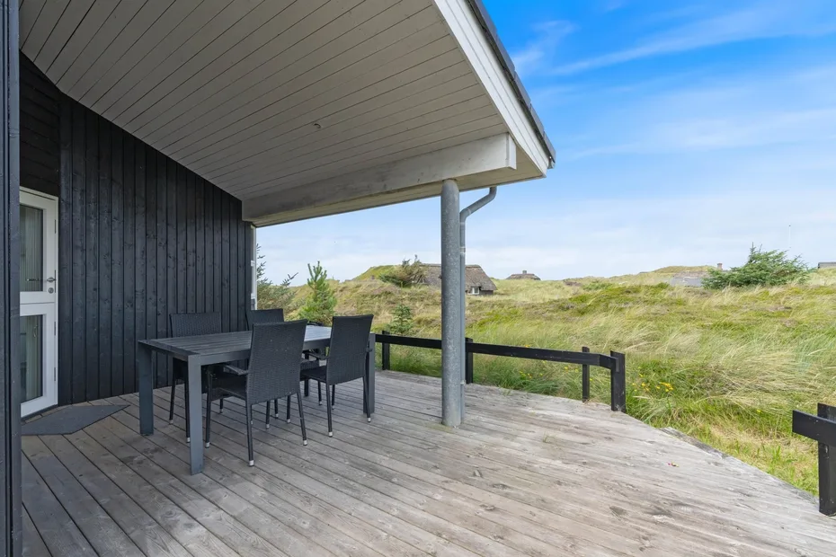 A wooden outdoor deck with a table and seating overlooks grassy dunes.