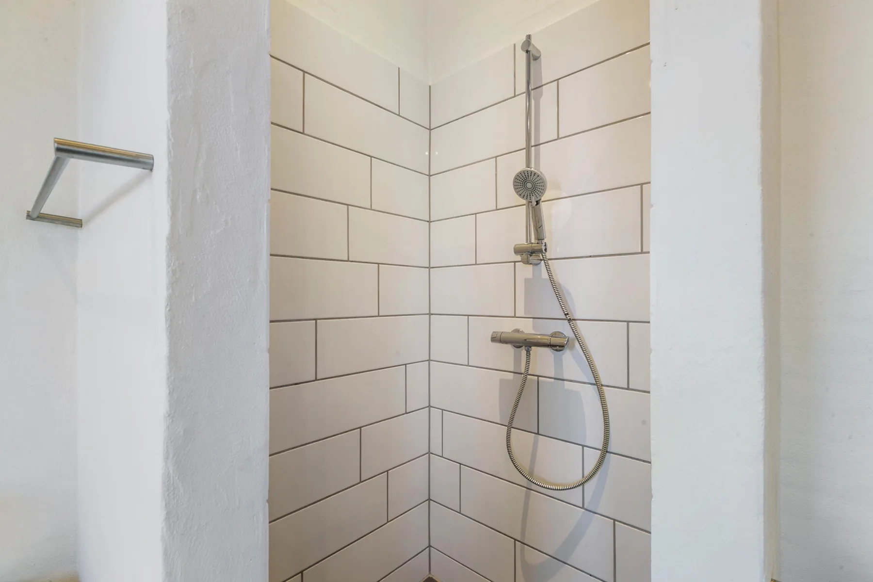 A tiled shower with a handheld showerhead and wall-mounted controls.