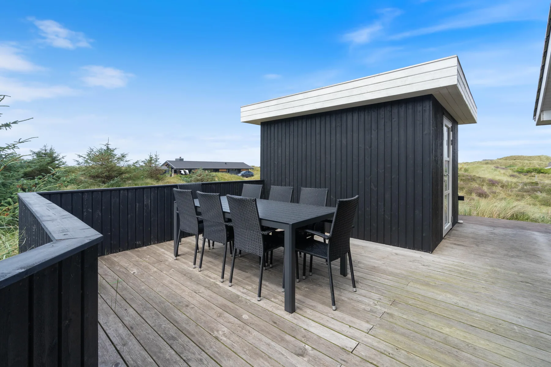 An outdoor deck has a dining table and seating beside a dark-sided building.