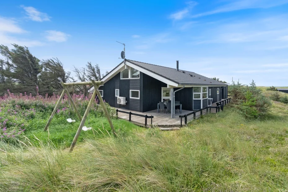 A dark modern house stands amid tall grass, with a small wooden swing nearby.