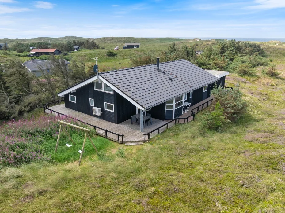 A dark house with a gray roof stands amid open grassy terrain.