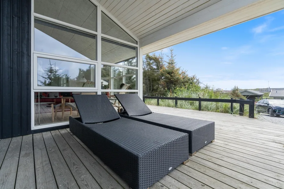 A covered outdoor deck with reclining lounge seating beside large windows.