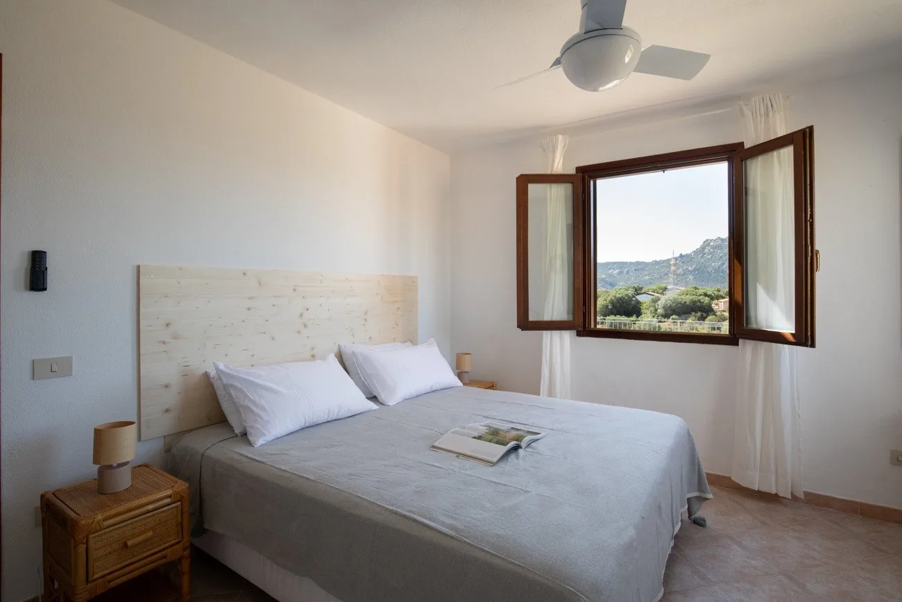 A bedroom with a bed beside an open window overlooking green hills.