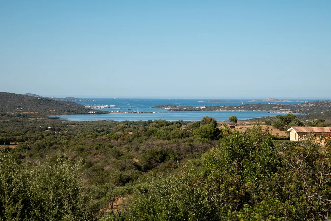 A broad coastal landscape with green hills and blue water under a clear sky.