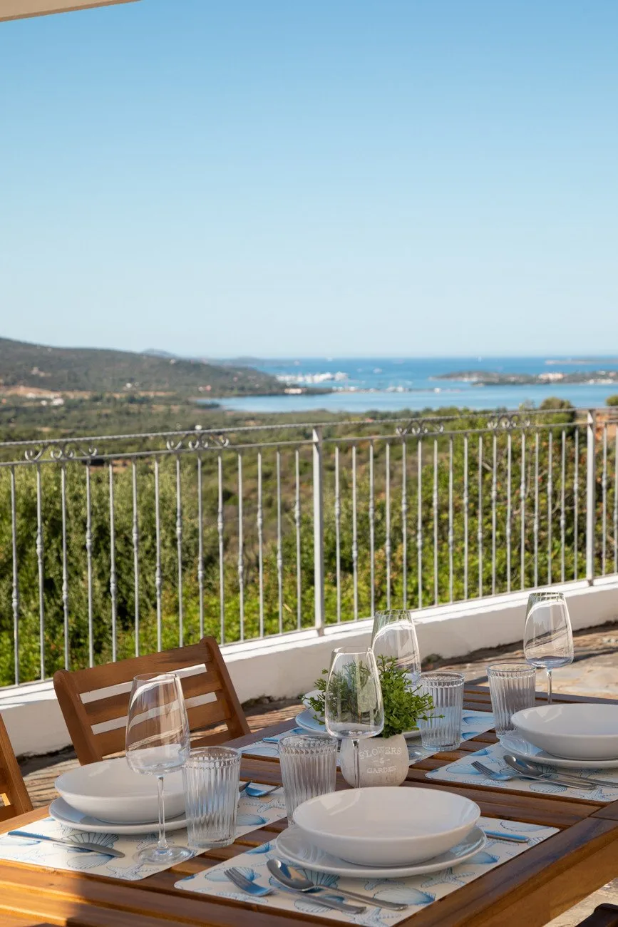 A terrace dining table overlooks green hills and a blue bay.