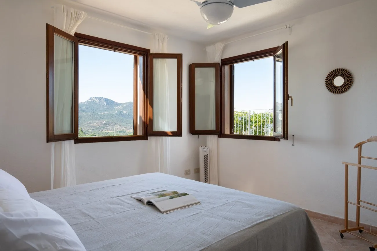 A bedroom with a neatly made bed and open windows overlooking hills.