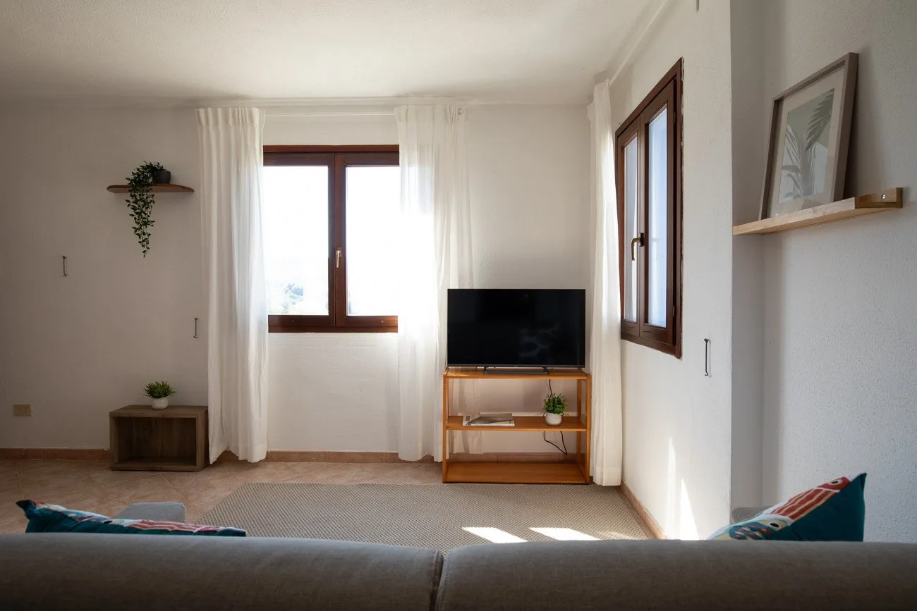 A bright living room with a television on a wooden stand beside two windows.