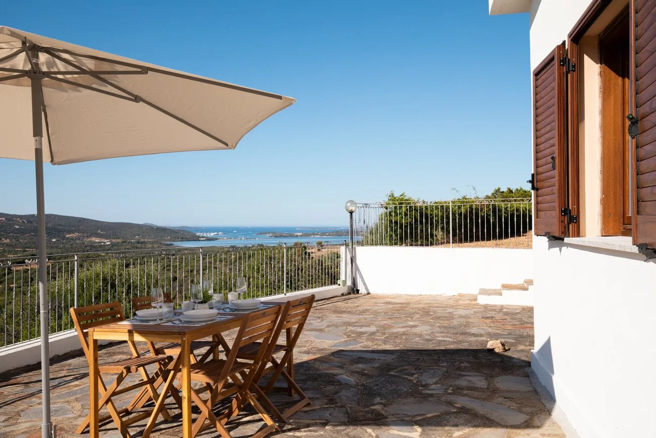 A terrace dining area with a wooden table, umbrella, and a view of the sea.