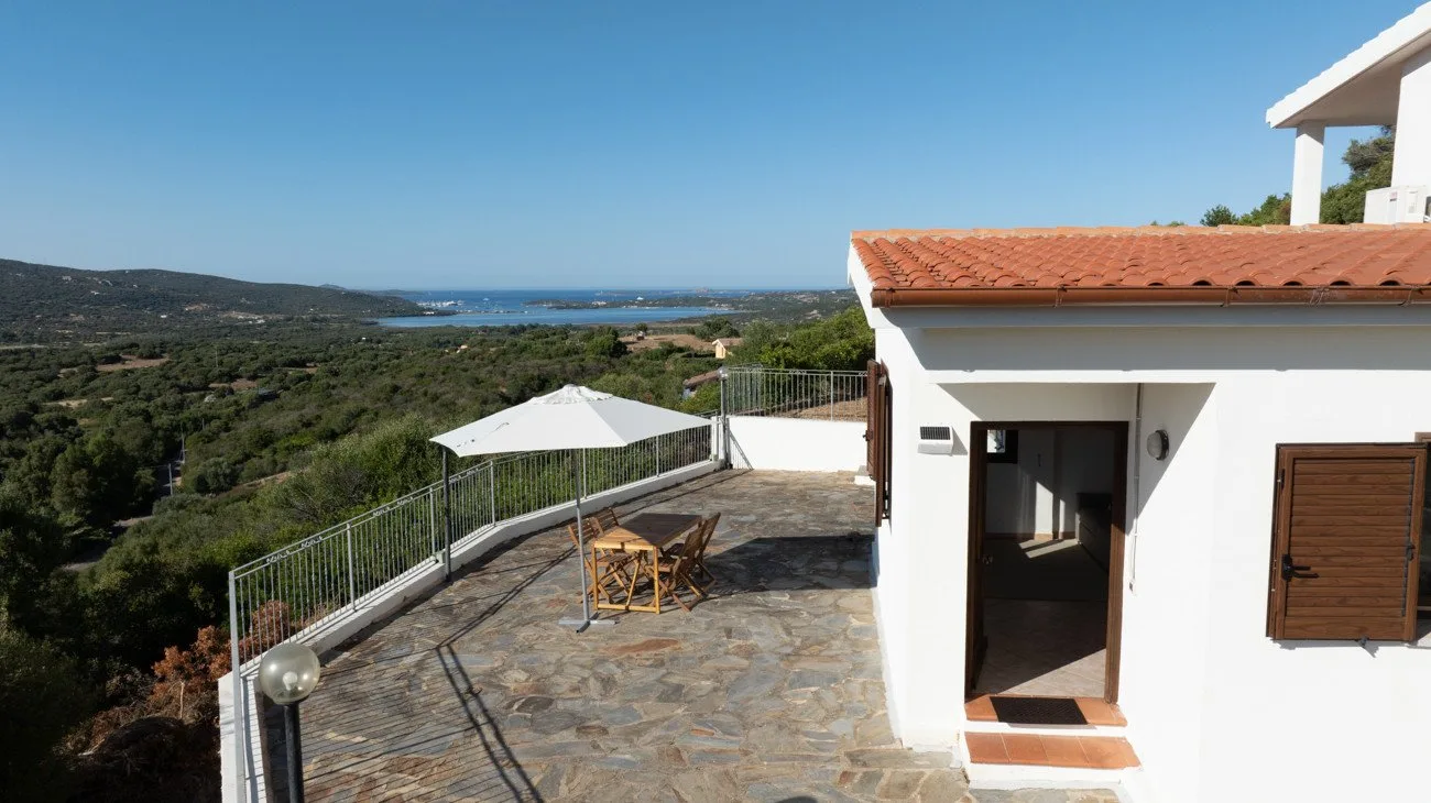 A stone terrace beside a white house overlooks green hills and the sea.