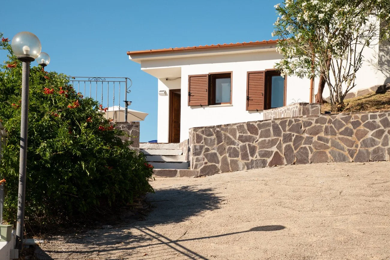 A white house with brown shutters stands above a stone retaining wall.
