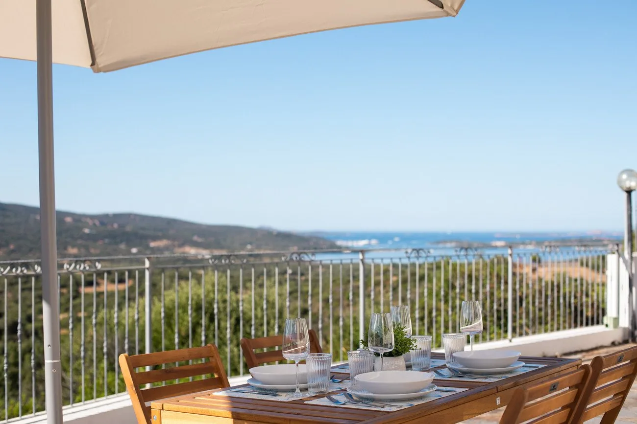 An outdoor dining table sits beneath an umbrella, overlooking a coastal landscape.