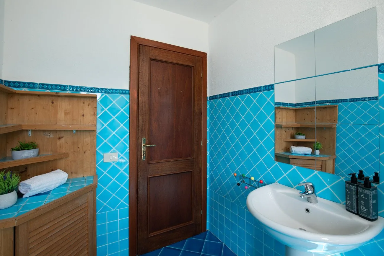 A bathroom with a wooden door, blue tiled walls, and a sink beneath a mirror.