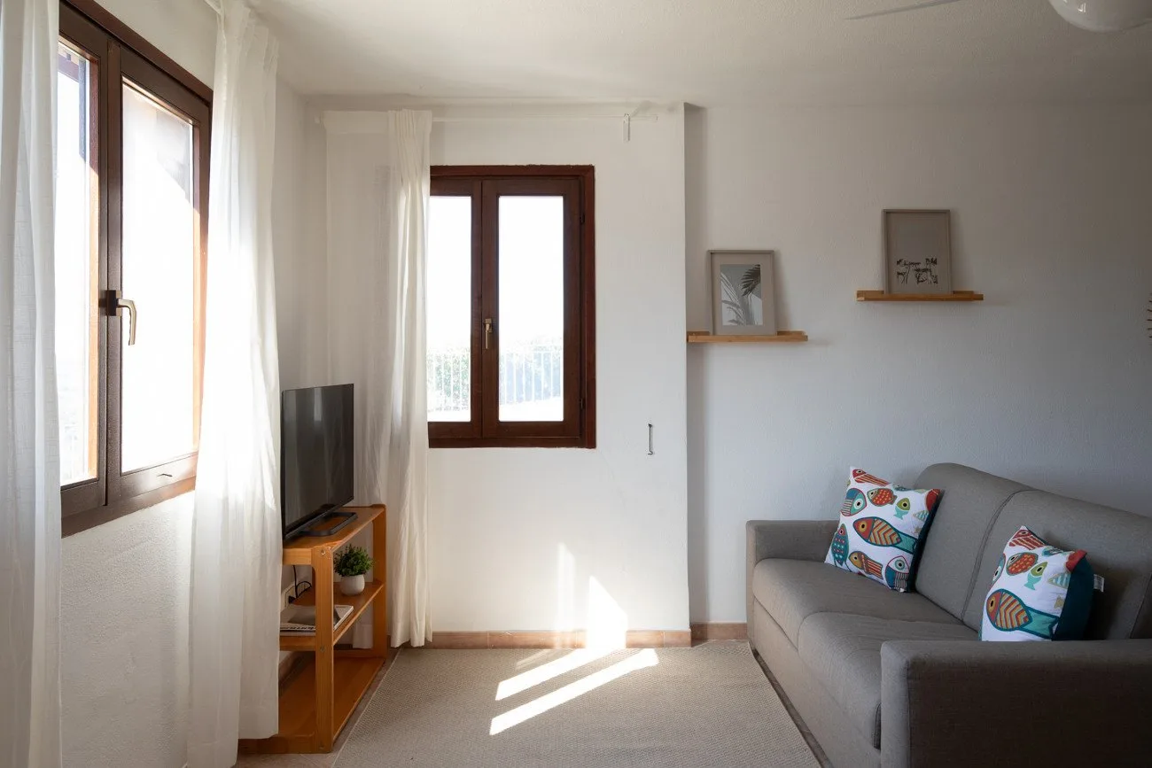 A bright living room with a gray sofa, television, and sunlight through the windows.