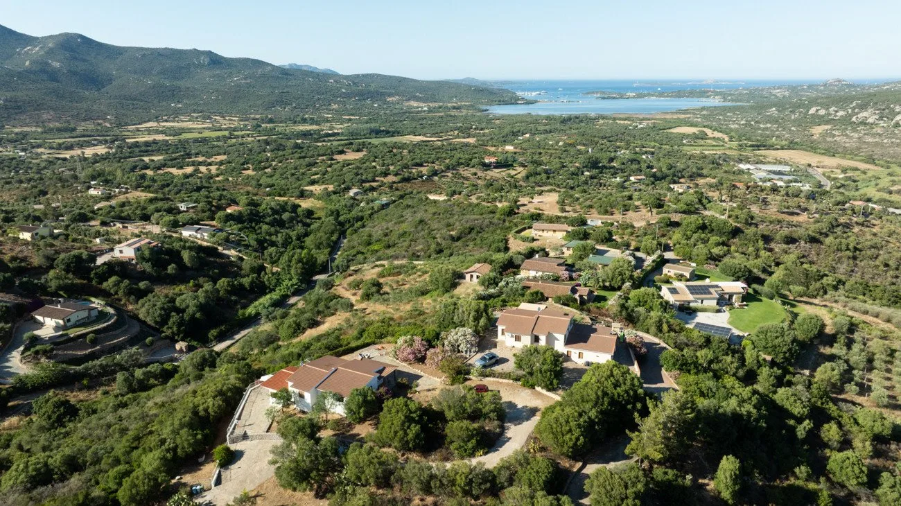 An aerial view of scattered homes among green hills, with water in the distance.
