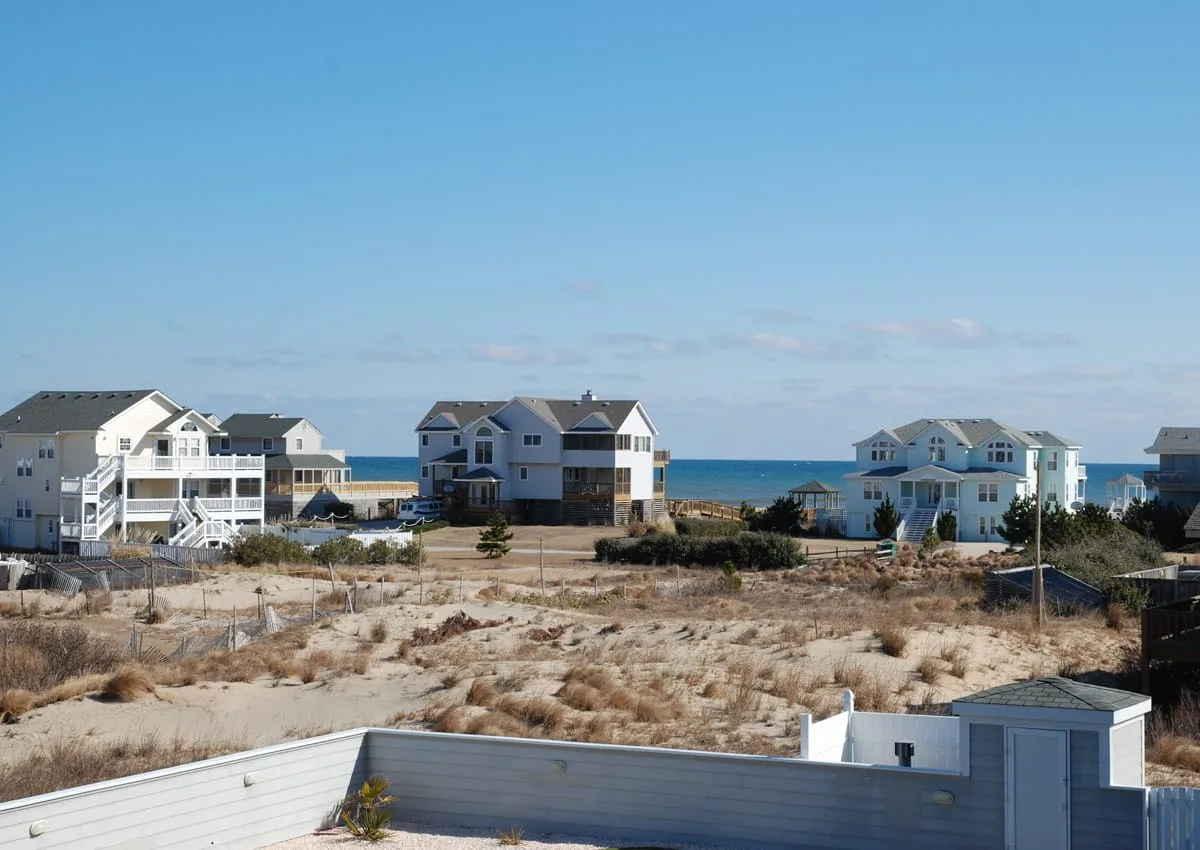 Coastal homes stand beyond sandy dunes, with the ocean visible in the background.