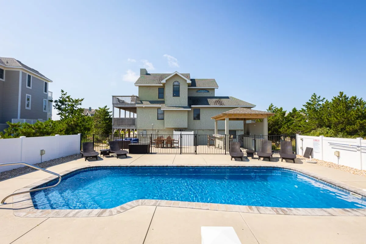 An outdoor swimming pool sits beside a large house under a clear sky.