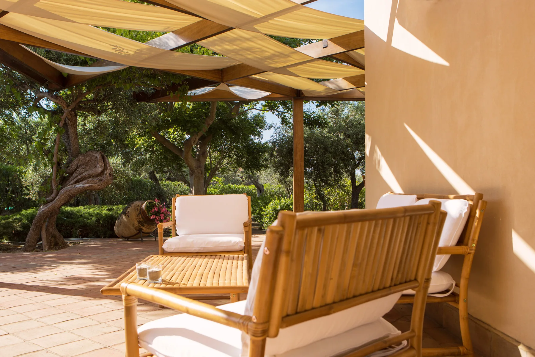 Un patio exterior con asientos de madera bajo una sombra de tela y árboles frondosos al fondo.