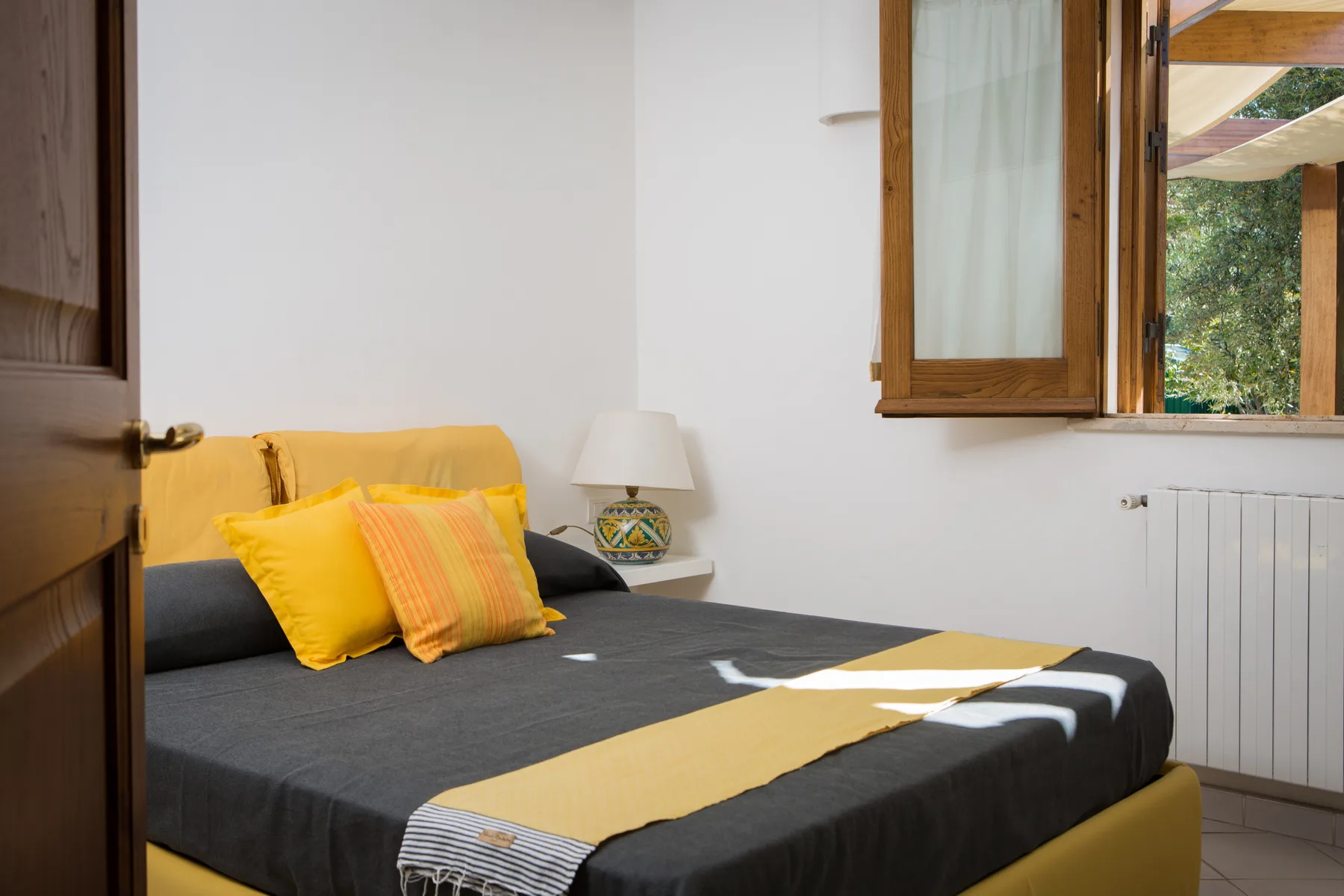 Un dormitorio con colcha oscura, almohadas amarillas y una ventana abierta.