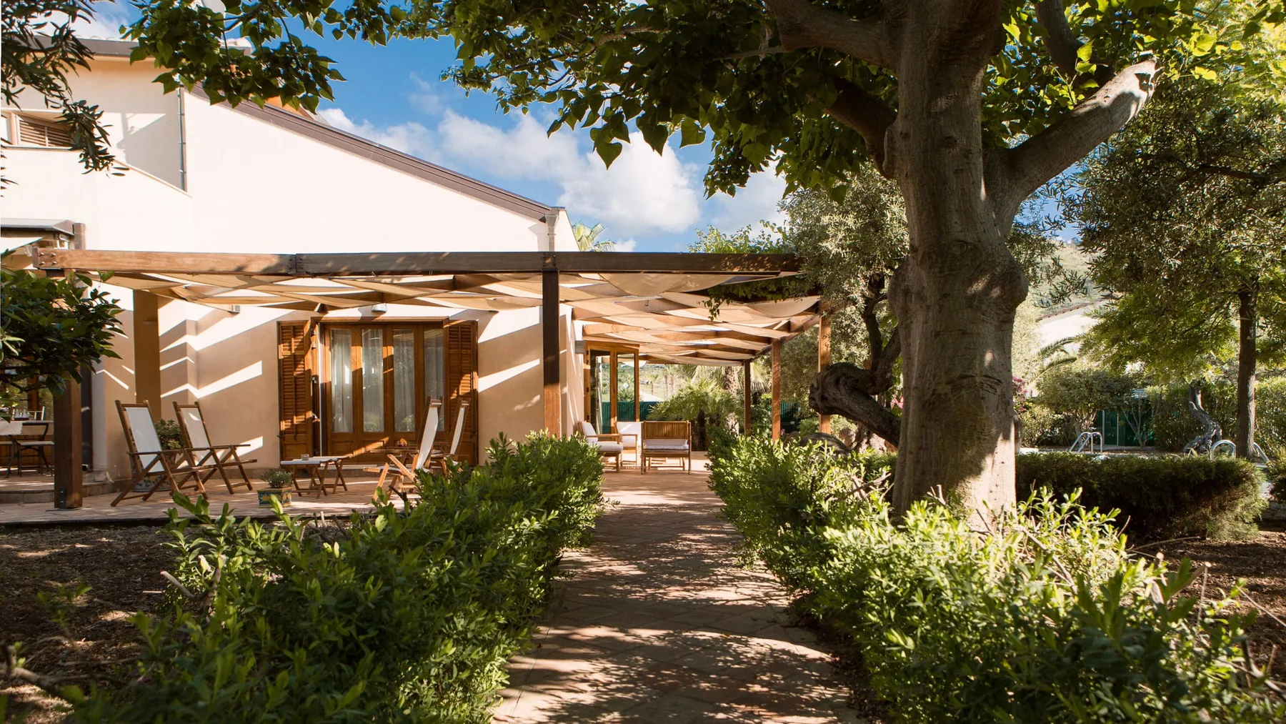 A shaded garden patio with a pergola, outdoor seating, and leafy trees.