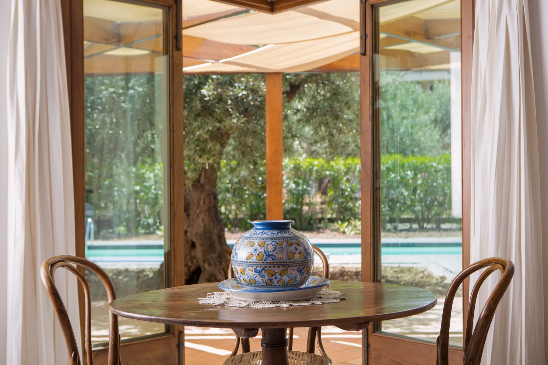 Una mesa redonda con un jarrón decorativo está junto a grandes ventanas con vista a una piscina.