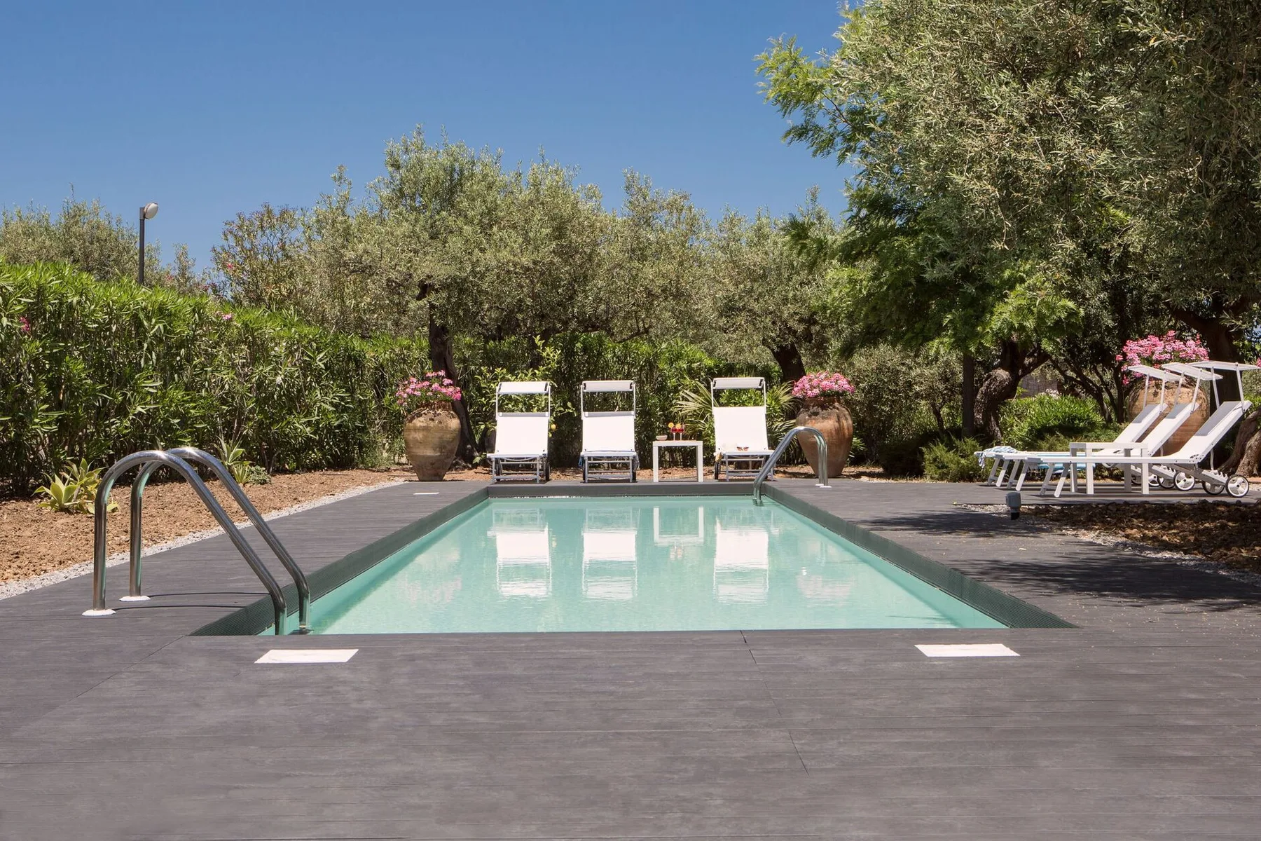 An outdoor swimming pool is surrounded by lounge chairs and leafy trees.