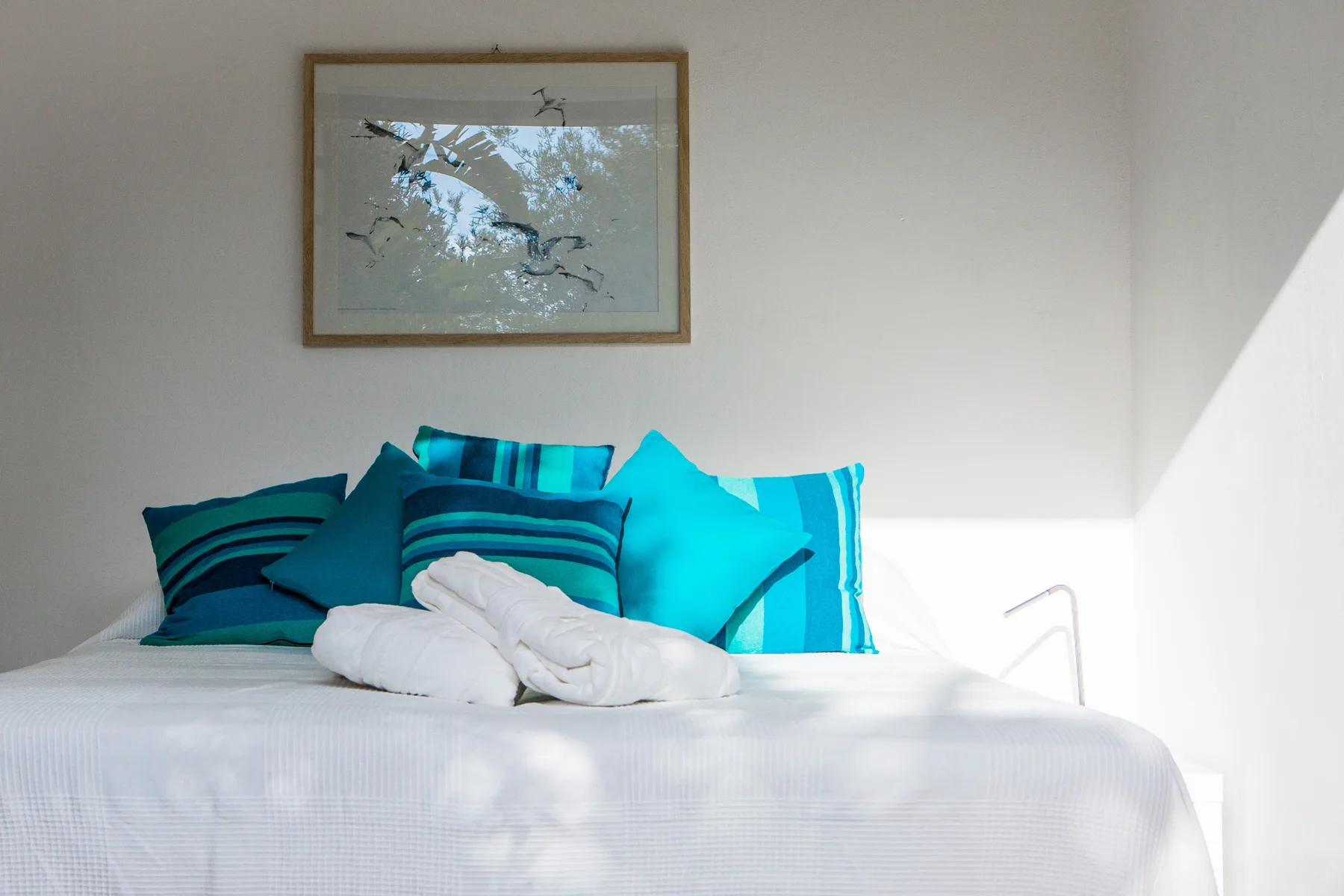Una cama bien arreglada con almohadas turquesas está bajo un cuadro enmarcado.
