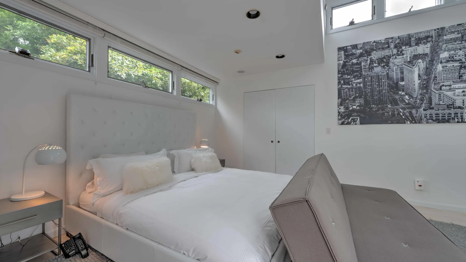 A bright bedroom with a bed beneath high windows and a large wall print.