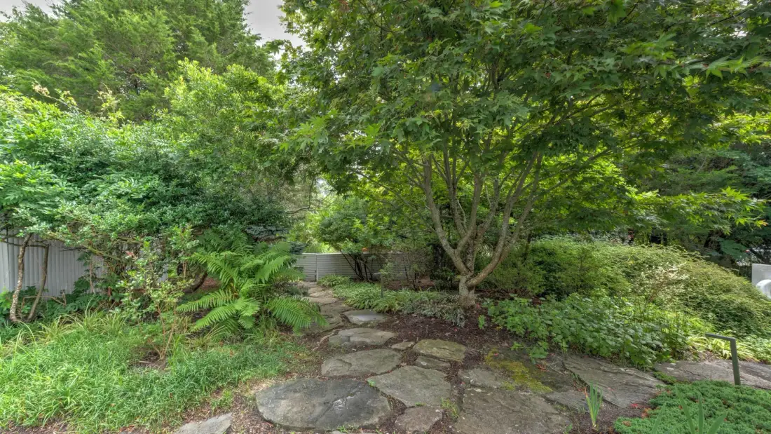 A stone path winds through a lush garden beneath leafy trees.