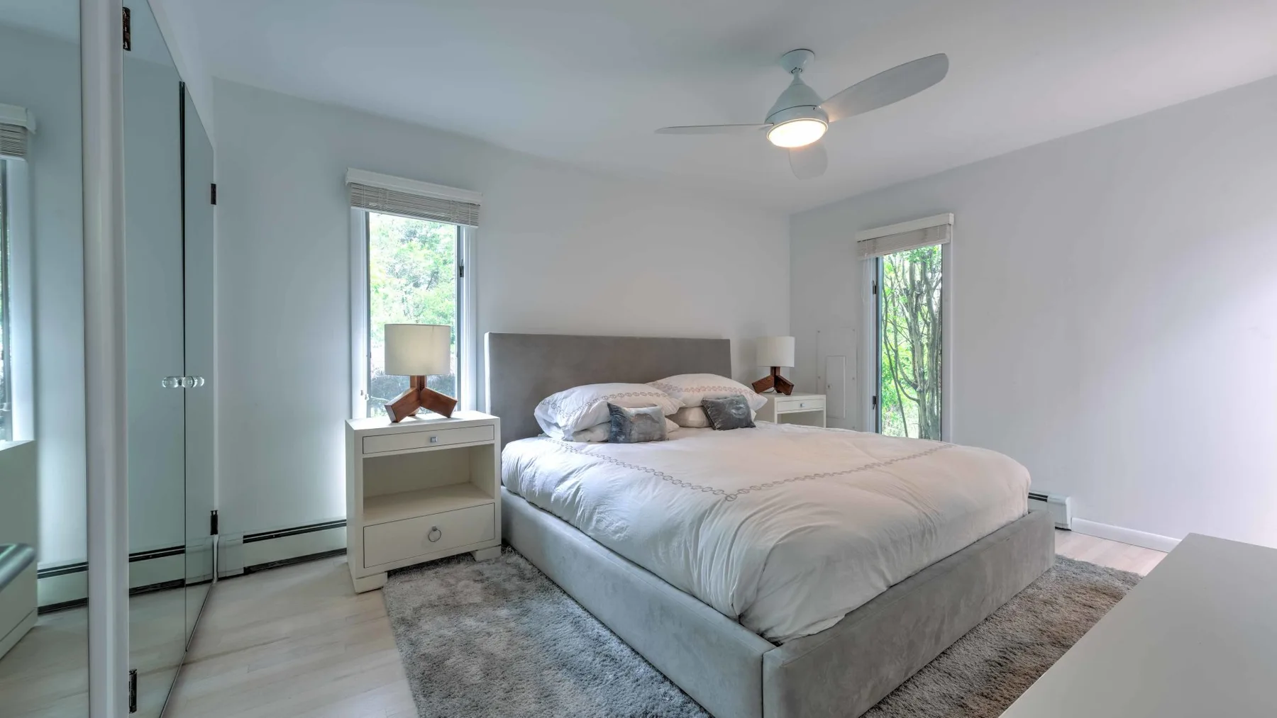 A bright bedroom with a neatly made bed and windows letting in daylight.