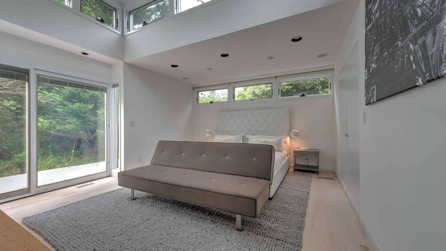 A bright bedroom with a bed, large windows, and a gray rug.