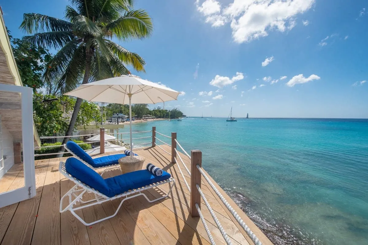 A seaside deck with blue loungers and an umbrella overlooks clear turquoise water.