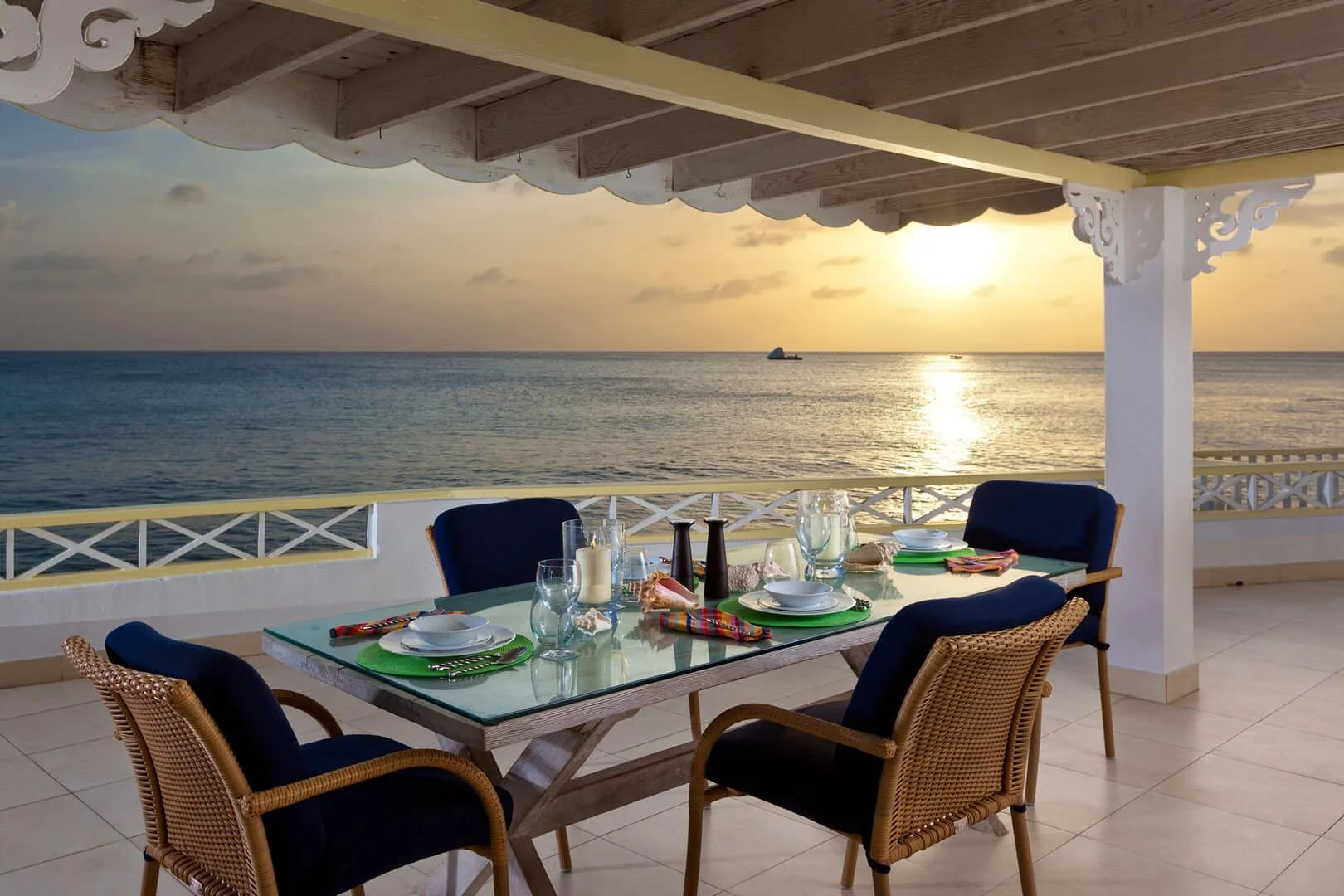 A seaside dining area with a table set beneath a covered terrace at sunset.
