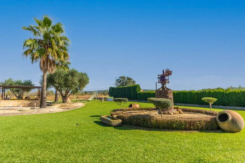 Un jardín paisajístico con una fuente central, setos recortados y palmeras.