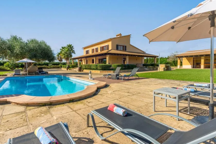Una piscina al aire libre junto a un patio con tumbonas y una sombrilla grande.