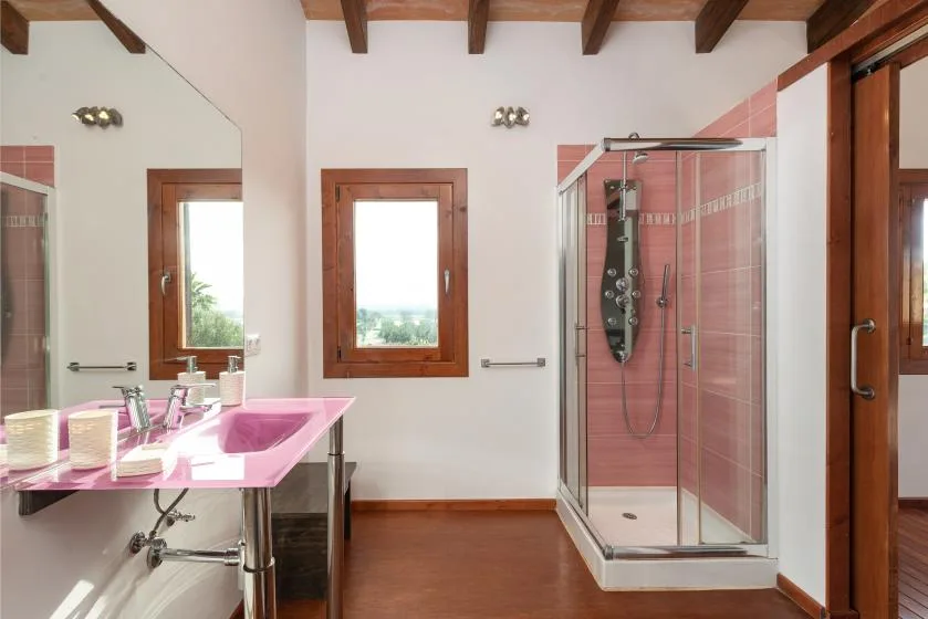 Un baño con lavabo rosa y ducha de vidrio junto a una ventana con marco de madera.
