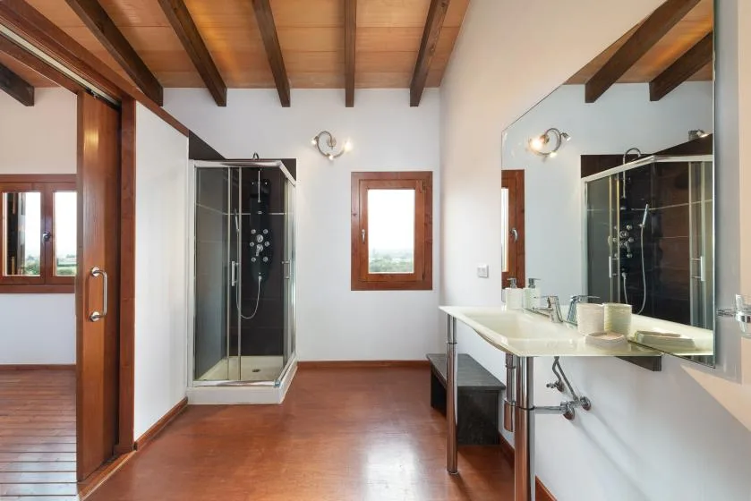 Un baño con ducha de vidrio, un lavabo alargado y vigas de madera a la vista.