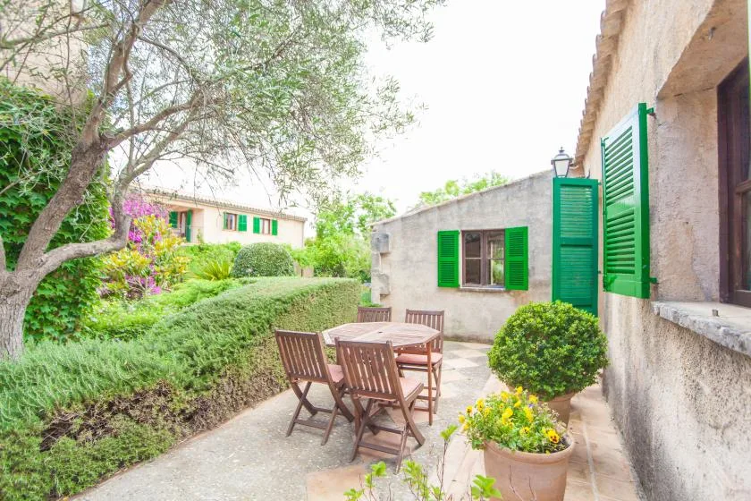 Un patio ajardinado con mesa y asientos de madera junto a una casa con contraventanas verdes.