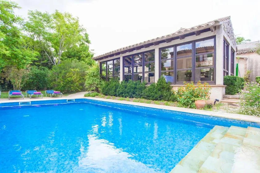 Una piscina está junto a un patio acristalado rodeado de vegetación.