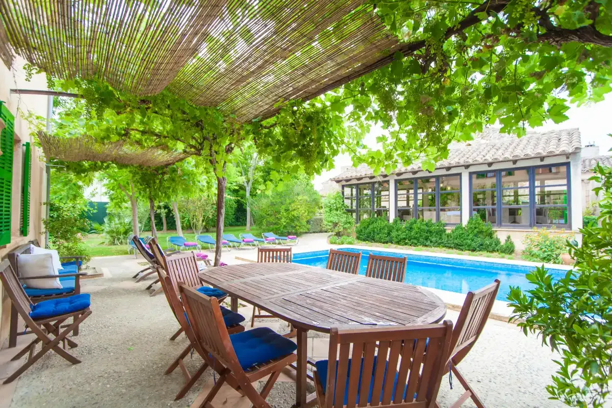 Una zona de comedor exterior a la sombra da a una piscina junto a una pérgola cubierta de enredaderas.